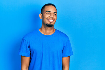 Young african american man wearing casual blue t shirt looking away to side with smile on face,...