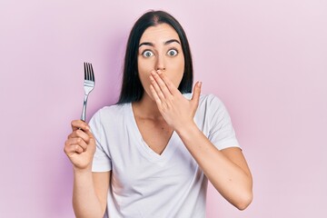 Beautiful woman with blue eyes holding one silver fork covering mouth with hand, shocked and afraid...