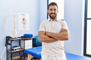 Young hispanic man wearing physiotherapist uniform standing with arms crossed gesture at clinic