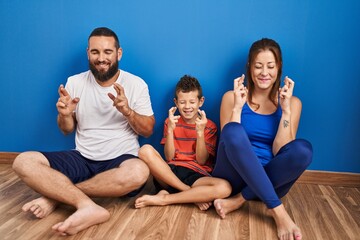 Family of three sitting on the floor at home gesturing finger crossed smiling with hope and eyes closed. luck and superstitious concept.