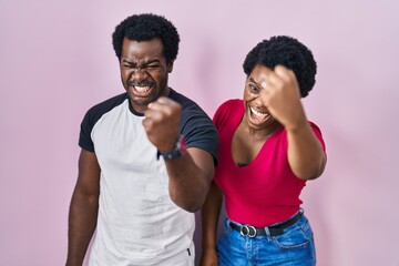 Young african american couple standing over pink background angry and mad raising fist frustrated and furious while shouting with anger. rage and aggressive concept.