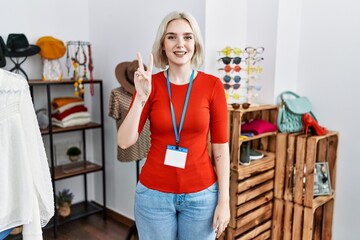 Young caucasian woman working as manager at retail boutique showing and pointing up with fingers number two while smiling confident and happy.