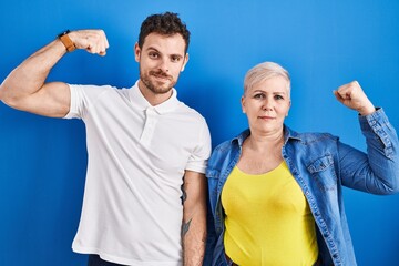 Young brazilian mother and son standing over blue background strong person showing arm muscle,...