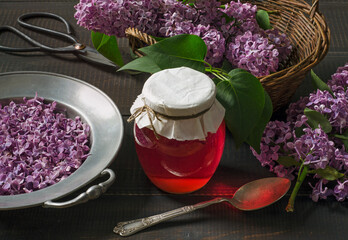Lilac flowers syrup in glass jar, lilac flowers and petals on dark wooden table.