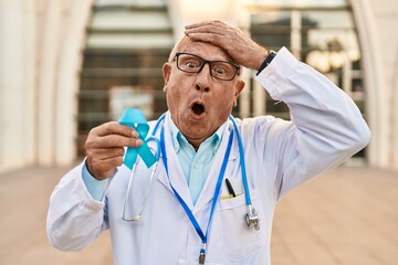 Senior doctor with grey hair holding blue ribbon stressed and frustrated with hand on head,...