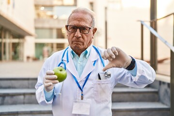 Senior doctor with grey hair holding healthy green apple with angry face, negative sign showing...