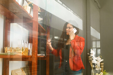 Latin American businesswoman holding a glass from a shelf of items and making a phone call with a white mobile.