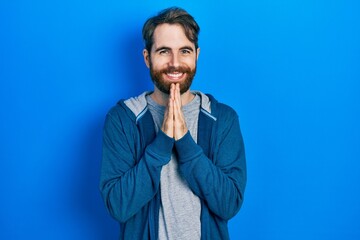 Caucasian man with beard wearing casual sweatshirt praying with hands together asking for forgiveness smiling confident.