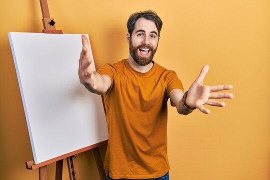 Caucasian man with beard standing by painter easel stand looking at the camera smiling with open arms for hug. cheerful expression embracing happiness.