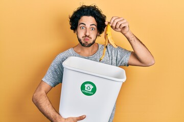 Handsome hispanic man recycling banana peel composting organic fertilizer puffing cheeks with funny face. mouth inflated with air, catching air. - obrazy, fototapety, plakaty