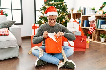 Young hispanic man unpacking gift sitting by christmas tree at home