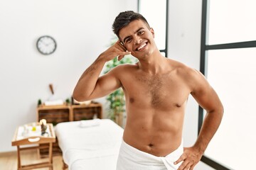 Young hispanic man standing shirtless at spa center smiling doing phone gesture with hand and fingers like talking on the telephone. communicating concepts.