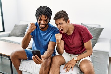 Young hispanic men using smartphone sitting on the sofa at home angry and mad raising fist...