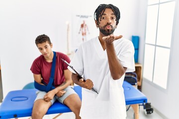Young hispanic man working at pain recovery clinic with a man with broken arm looking at the camera blowing a kiss with hand on air being lovely and sexy. love expression.