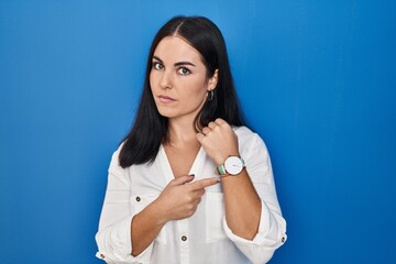 Young hispanic woman standing over blue background in hurry pointing to watch time, impatience,...