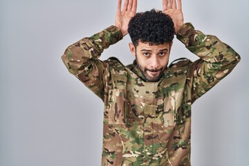 Arab man wearing camouflage army uniform doing bunny ears gesture with hands palms looking cynical and skeptical. easter rabbit concept.
