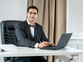 A businessman looks at the camera with a smile. The man works in the office for a laptop. A business person looks welcome. The man works at the table.