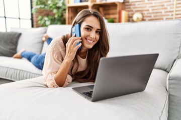 Naklejka premium Young hispanic woman smiling confident using laptop and talking on the smartphone at home