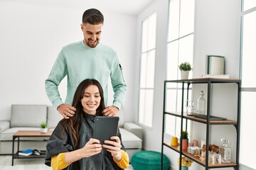 Obraz premium Young hispanic woman looking touchapd screen while stylist man comb hair at beauty center.
