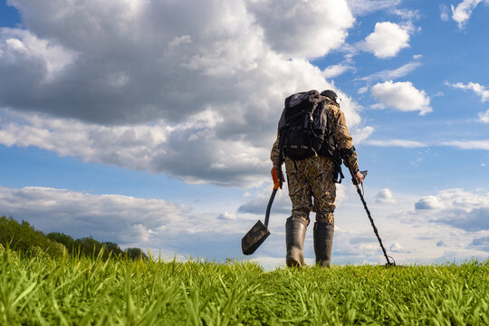 Man with metal detector with his back to camera. Man in military clothes with metal detector. Digger in summer meadow. Concept searching for weapons underground. Digger with metal detector