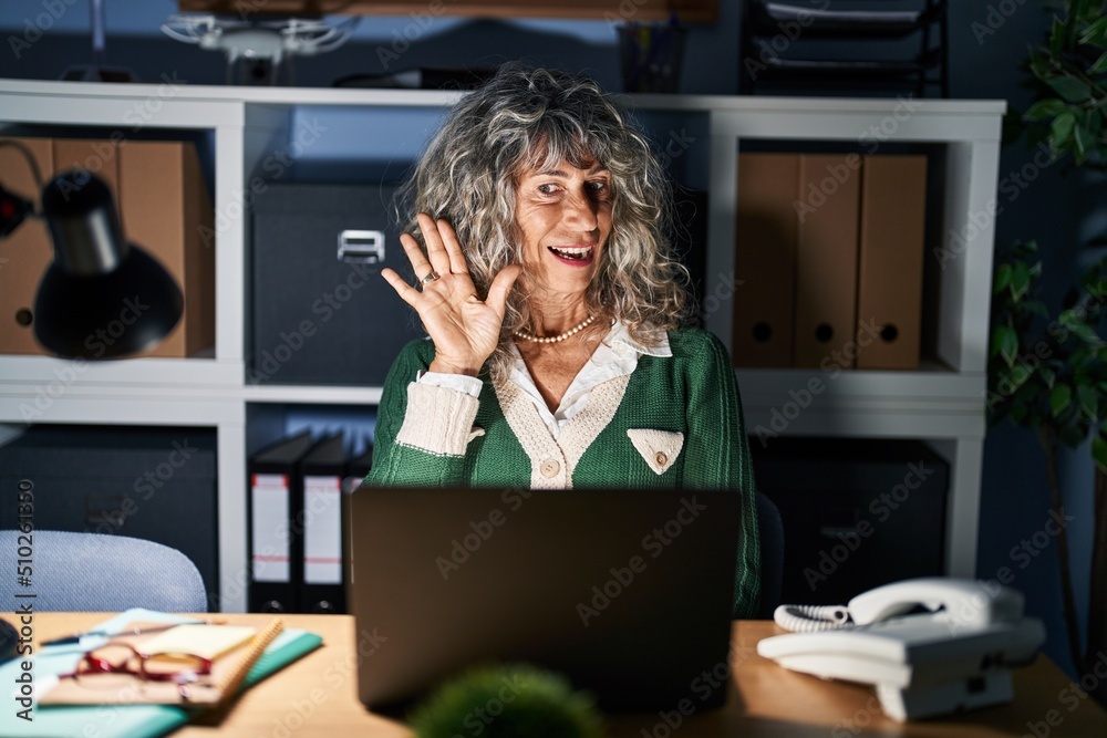 Wall mural Middle age woman working at night using computer laptop waiving saying hello happy and smiling, friendly welcome gesture