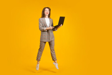 Girl with laptop. Woman is typing on computer. Woman office worker in full growth. Businesswoman standing with laptop. Concept girl works on Internet. Businesswoman in plaid suit on yellow