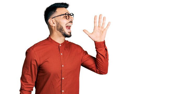 Young hispanic man with beard wearing business shirt and glasses shouting and screaming loud to side with hand on mouth. communication concept.