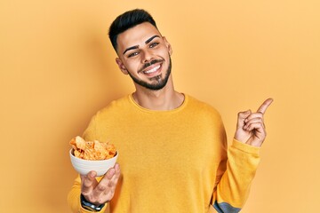Young hispanic man with beard holding nachos potato chips smiling happy pointing with hand and finger to the side