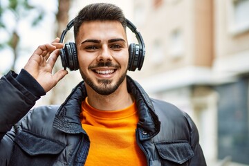 Handsome hispanic man with beard smiling happy and confident at the city wearing winter coat and headphones