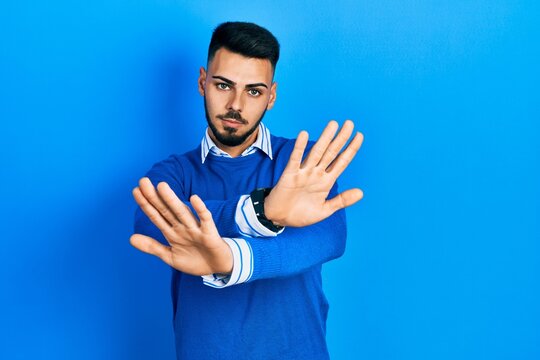 Young hispanic man with beard wearing casual blue sweater rejection expression crossing arms doing negative sign, angry face