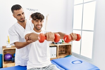 Two hispanic men physiotherapist and patient having rehab session using dumbbells at clinic