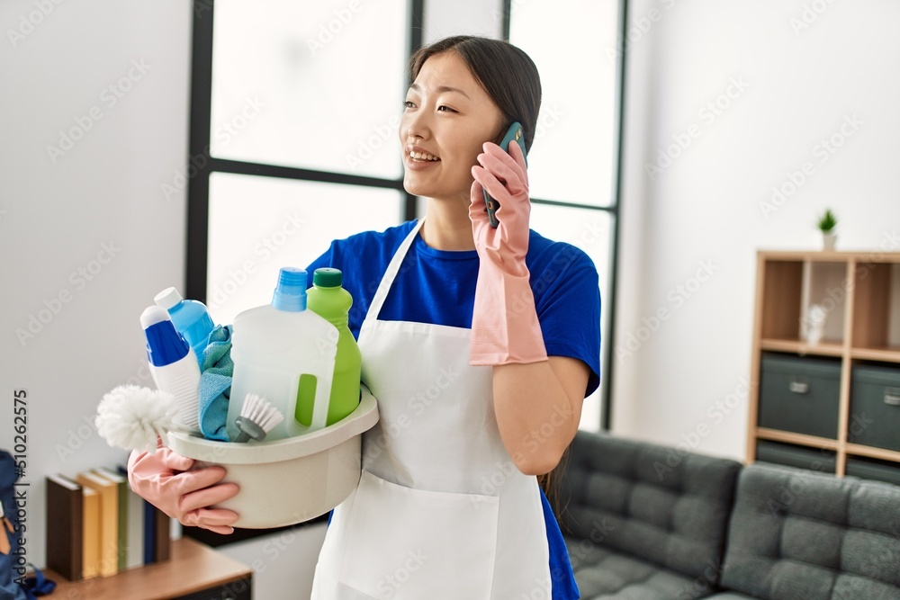 Wall mural young chinese housewife holding cleaning products talking on the smartphone at home.