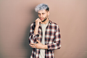 Young hispanic man with modern dyed hair wearing casual shirt serious face thinking about question with hand on chin, thoughtful about confusing idea