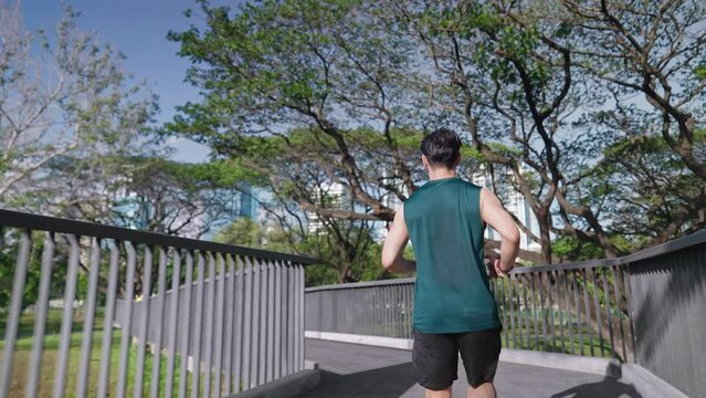 Shot from behind of light skin Asian man running on the walking track inside recreation park on hot and sunny day, forest inside the city downtown, outdoor cardio exercise, jogging man in sport wear