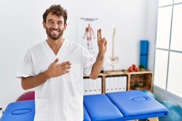 Young handsome physiotherapist man working at pain recovery clinic smiling swearing with hand on...