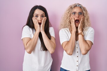 Mother and daughter standing together over pink background afraid and shocked, surprise and amazed expression with hands on face