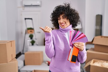 Young brunette woman with curly hair choosing color of new house wall pointing thumb up to the side...