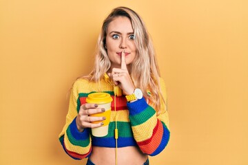 Beautiful young blonde woman drinking cup of coffee wearing headphones asking to be quiet with finger on lips. silence and secret concept.