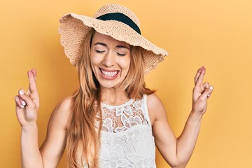 Young caucasian woman wearing summer hat gesturing finger crossed smiling with hope and eyes closed. luck and superstitious concept.