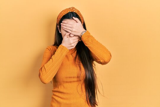 Young brunette teenager wearing casual yellow sweater covering eyes and mouth with hands, surprised and shocked. hiding emotion