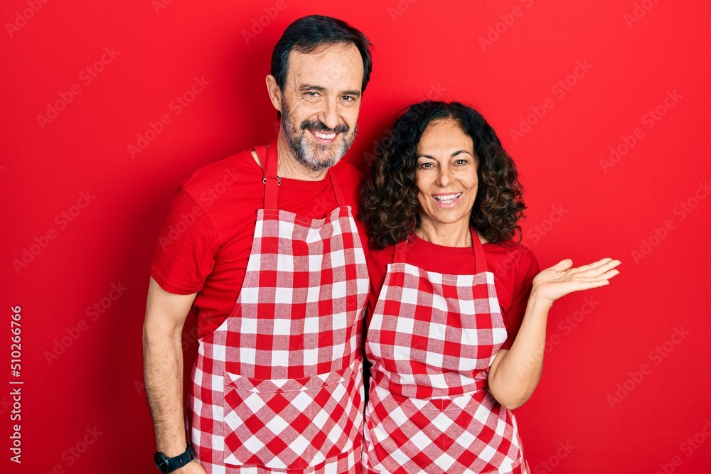 Sticker Middle age couple of hispanic woman and man wearing cook apron smiling cheerful presenting and pointing with palm of hand looking at the camera.