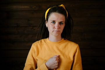 Young sad woman in yellow hoodie with dreadlocks holds one hand in fist near heart at chest....
