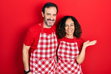 Middle age couple of hispanic woman and man wearing cook apron smiling cheerful presenting and pointing with palm of hand looking at the camera.