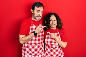 Middle age couple of hispanic woman and man wearing cook apron pointing aside worried and nervous with forefinger, concerned and surprised expression