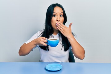 Beautiful hispanic woman with nose piercing drinking a cup coffee covering mouth with hand, shocked and afraid for mistake. surprised expression