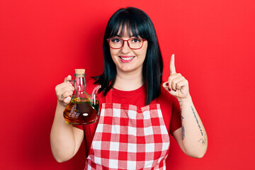 Young hispanic woman wearing cook apron holding olive oil can smiling with an idea or question...