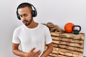 African american man listening to music using headphones at the gym with hand on stomach because nausea, painful disease feeling unwell. ache concept.