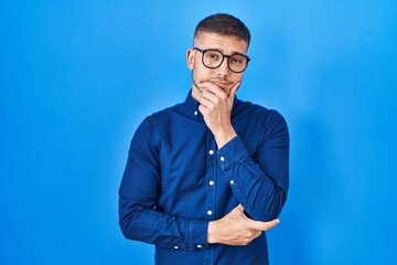 Young hispanic man wearing glasses over blue background looking confident at the camera smiling with crossed arms and hand raised on chin. thinking positive.