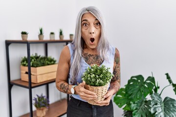 Middle age grey-haired woman holding green plant pot at home in shock face, looking skeptical and sarcastic, surprised with open mouth