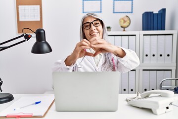 Middle age grey-haired woman wearing doctor uniform working using computer laptop smiling in love showing heart symbol and shape with hands. romantic concept.